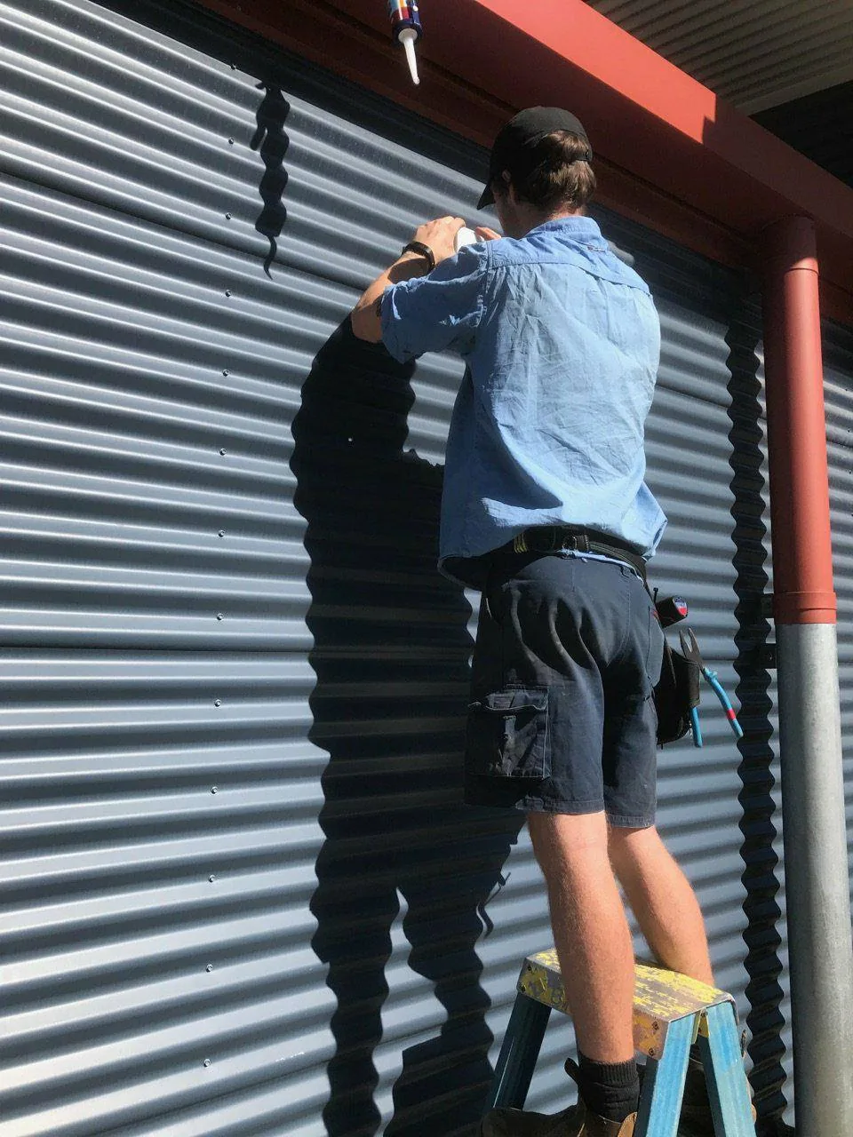Electrician on a step ladder working on the wall beside a roller door, seen from behind