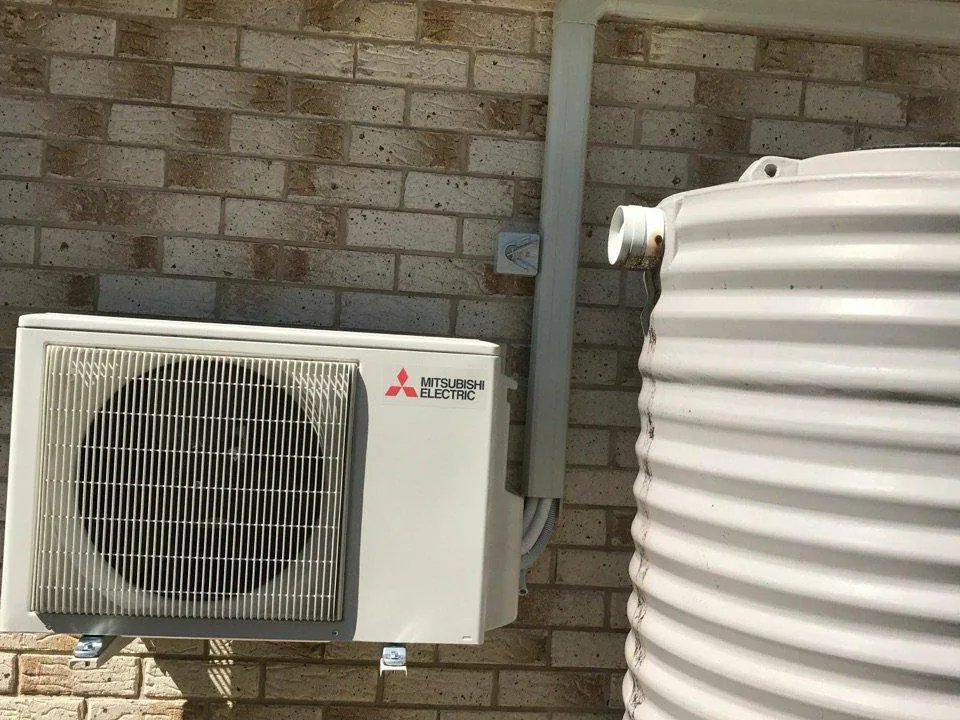 New Mitsubishi Electric split system outdoor unit mounted on a brick wall beside a rainwater tank, with neat white ducting